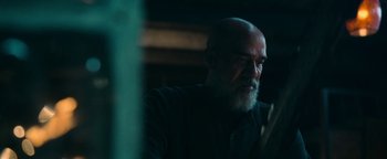 Movie still from “No One Gets Out Alive” (2021), directed by Santiago Menghini – An older man with a beard is sitting in a dark room; Close Up shot, Over the shoulder angle
