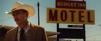Movie still from “Nocturnal Animals” (2016), directed by Tom Ford – A man in a suit and hat standing in front of a motel sign; Medium shot, Low angle