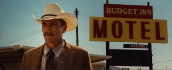Movie still from “Nocturnal Animals” (2016), directed by Tom Ford – A man wearing a cowboy hat standing in front of a motel sign; Medium shot, Low angle