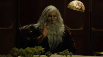Movie still from “Oats Studios” (2017), directed by Neill Blomkamp – An old man with long white hair and a long white beard sitting at a table; Medium shot, Over the shoulder angle
