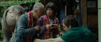Movie still from “Okja” (2017), directed by Bong Joon Ho – A young girl is looking at a book with an older man and woman; Medium shot, Over the shoulder angle