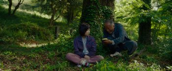 Movie still from “Okja” (2017), directed by Bong Joon Ho – An older man and a young woman sitting in the grass; Medium shot, High angle