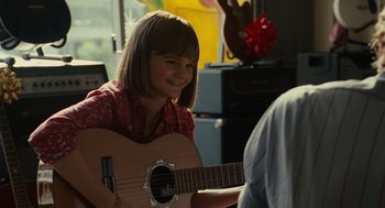 Movie still from “Moneyball” (2011), directed by Bennett Miller – A young girl is playing a guitar in a room; Medium shot, Over the shoulder angle