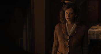 Movie still from “Operation Finale” (2018), directed by Chris Weitz – A woman standing in a room wearing a brown jacket; Medium shot, Over the shoulder angle