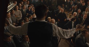 Movie still from “Operation Finale” (2018), directed by Chris Weitz – A crowd of people sitting and standing in a room; Medium shot, Over the shoulder angle