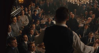 Movie still from “Operation Finale” (2018), directed by Chris Weitz – A crowd of people sitting in a room with a man in a vest; Wide shot, High angle