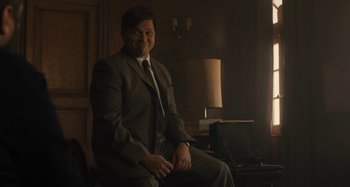 Movie still from “Operation Finale” (2018), directed by Chris Weitz – A man in a suit and tie sitting in a chair; Medium shot, Over the shoulder angle