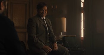 Movie still from “Operation Finale” (2018), directed by Chris Weitz – A man in a suit sitting in a chair in front of a lamp; Medium shot, Over the shoulder angle