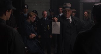 Movie still from “Operation Finale” (2018), directed by Chris Weitz – A man in a suit and hat holding up a drawing; Medium shot, Over the shoulder angle