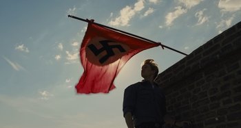 Movie still from “Operation Finale” (2018), directed by Chris Weitz – A man standing next to a red and black flag; Medium shot, Low angle