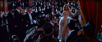Movie still from “Moulin Rouge!” (2001), directed by Baz Luhrmann – A crowd of people sitting and standing around a woman in a white dress; Wide shot, High angle
