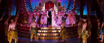 Movie still from “Moulin Rouge!” (2001), directed by Baz Luhrmann – A couple is standing on a stage with a bunch of dancers; Wide shot, Over the shoulder angle