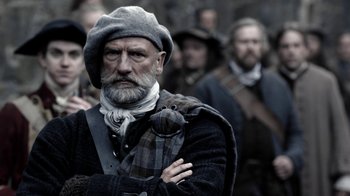 Movie still from “Outlander” (2014), created by Ronald D. Moore – An old man with a beard wearing a hat and holding a bird; Medium shot, Low angle