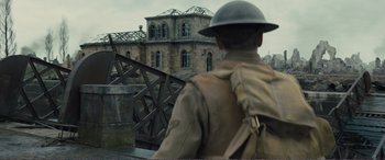 Movie still from “1917” (2019), directed by Sam Mendes – A man wearing a hat and a backpack looking at a building; Medium shot, Over the shoulder angle