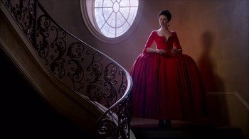 Movie still from “Outlander” (2014), created by Ronald D. Moore – A woman in a red dress standing in front of a staircase; Wide shot, Low angle