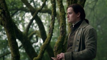 Movie still from “Outlander” (2014), created by Ronald D. Moore – A man standing next to a tree in the woods; Medium shot, Low angle