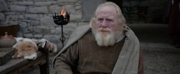 Movie still from “Outlaw King” (2018), directed by David Mackenzie – An old man with a white beard sitting next to a fire pit; Medium shot, Low angle