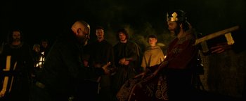Movie still from “Outlaw King” (2018), directed by David Mackenzie – A group of people standing around a fire; Medium shot, Over the shoulder angle