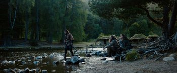 Movie still from “Outlaw King” (2018), directed by David Mackenzie – A group of men sitting on rocks near a body of water; Wide shot, High angle