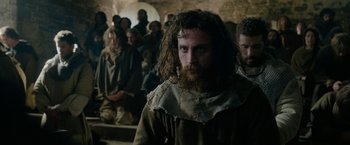 Movie still from “Outlaw King” (2018), directed by David Mackenzie – A man with long hair and a long beard is standing in front of a group of people; Medium shot, Over the shoulder angle