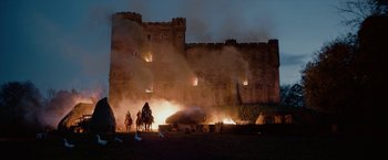 Movie still from “Outlaw King” (2018), directed by David Mackenzie – A group of people riding horses next to a burning building; Extreme Wide shot, Low angle