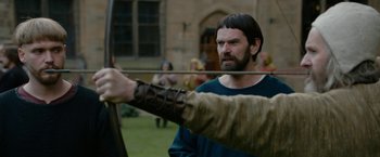 Movie still from “Outlaw King” (2018), directed by David Mackenzie – A man with long hair and a beard is holding a bow and arrow; Medium shot, Over the shoulder angle