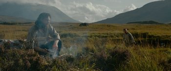 Movie still from “Outlaw King” (2018), directed by David Mackenzie – A person sitting in a field near a fire; Wide shot, Low angle