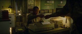 Movie still from “Outside the Wire” (2021), directed by Mikael Håfström – A man sitting in front of a printer holding his hands; Medium shot, Low angle