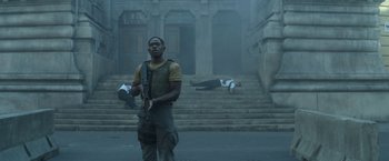 Movie still from “Outside the Wire” (2021), directed by Mikael Håfström – A man holding a gun while standing in front of a building; Medium shot, Low angle