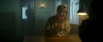 Movie still from “Outside the Wire” (2021), directed by Mikael Håfström – A woman sitting at a wooden table in a dark room; Medium shot, Low angle