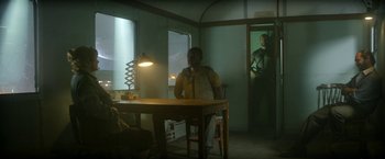 Movie still from “Outside the Wire” (2021), directed by Mikael Håfström – A man standing in a dimly - lit room next to a table; Wide shot, Over the shoulder angle