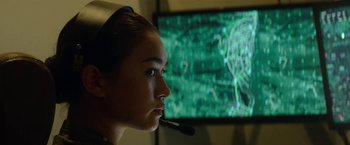 Movie still from “Outside the Wire” (2021), directed by Mikael Håfström – A woman with a headset in front of a television; Close Up shot, Low angle