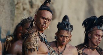 Movie still from “Apocalypto” (2006), directed by Mel Gibson – A man in a native american costume with other men in the background; Close Up shot, Low angle
