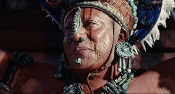 Movie still from “Apocalypto” (2006), directed by Mel Gibson – An older man wearing a headdress with beads around his neck; Close Up shot, Low angle