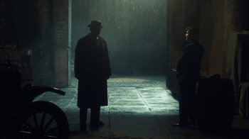 Movie still from “Peaky Blinders” (2013), created by Steven Knight – A man wearing a hat and a long coat standing in the rain with another man; Wide shot, Over the shoulder angle