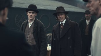 Movie still from “Peaky Blinders” (2013), created by Steven Knight – A couple of men standing next to each other wearing hats; Medium shot, Over the shoulder angle