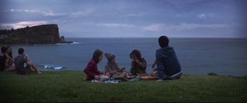 Movie still from “Penguin Bloom” (2020), directed by Glendyn Ivin – A group of people sitting in the grass near the ocean; Wide shot, High angle