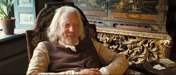 Movie still from “Pride & Prejudice” (2005), directed by Joe Wright – An older man sitting in a chair in front of an oriental painting; Medium shot, High angle