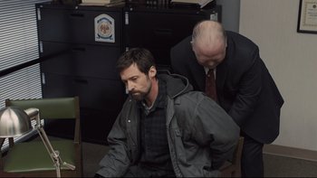 Movie still from “Prisoners” (2013), directed by Denis Villeneuve – A man sitting on a chair in a room with another man standing next to him; Medium shot, High angle