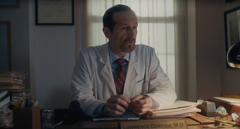 Movie still from “Private Life” (2018), directed by Tamara Jenkins – A man sitting at a desk wearing a white lab coat; Medium shot, Over the shoulder angle