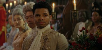 Movie still from “Queen Charlotte: A Bridgerton Story” (2023), directed by Tom Verica – A man wearing a suit and tie standing in front of candles; Close Up shot, Over the shoulder angle