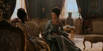 Movie still from “Queen Charlotte: A Bridgerton Story” (2023), directed by Tom Verica – A woman in a dress sitting on a chair in a room; Medium shot, Over the shoulder angle