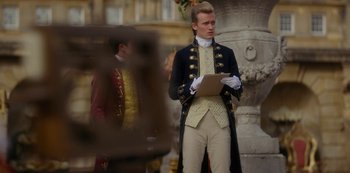 Movie still from “Queen Charlotte: A Bridgerton Story” (2023), directed by Tom Verica – A man in a suit holding a clipboard in front of a statue; Medium shot, Over the shoulder angle