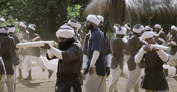 Movie still from “RRR” (2022), directed by S.S. Rajamouli – A group of men standing next to each other on a dirt field; Medium shot, Over the shoulder angle