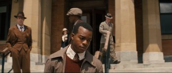 Movie still from “Race” (2016), directed by Stephen Hopkins – A man in a suit and tie standing in front of a building; Medium shot, Over the shoulder angle
