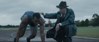 Movie still from “Race” (2016), directed by Stephen Hopkins – A man in a hat and a man in a trench coat and shorts; Medium shot, Over the shoulder angle