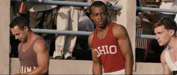 Movie still from “Race” (2016), directed by Stephen Hopkins – A man standing in front of a group of people; Medium shot, Over the shoulder angle