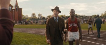 Movie still from “Race” (2016), directed by Stephen Hopkins – A man in a suit and tie next to another man in a red shirt and white shorts; Medium shot, Over the shoulder angle