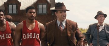 Movie still from “Race” (2016), directed by Stephen Hopkins – A man wearing a suit and hat next to another man; Medium shot, Over the shoulder angle