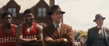 Movie still from “Race” (2016), directed by Stephen Hopkins – A man in a suit and hat standing in front of a building; Medium shot, Over the shoulder angle